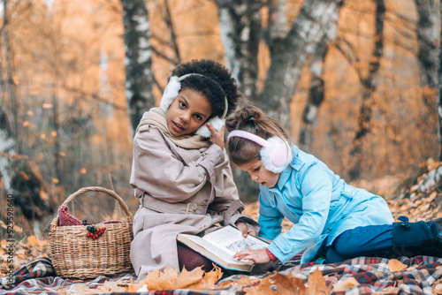 Happy Caucasian and African-American girls read a book together at a picnic in the autumn park.Diverse people autumn concept.