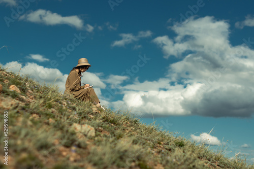 Side view of a woman in a hat sitting on the edge of a hill against a blue sky, drawing in her sketchbook