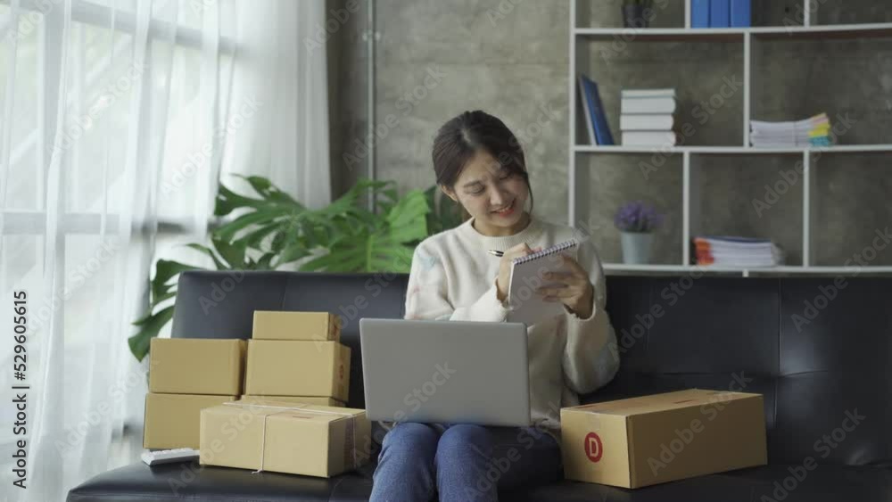 Asian woman sitting on sofa at home with box and laptop taking orders from customers online business sme sales concept Asian woman sitting on sofa at home with box and laptop taking orders from customers online business sme sales concept