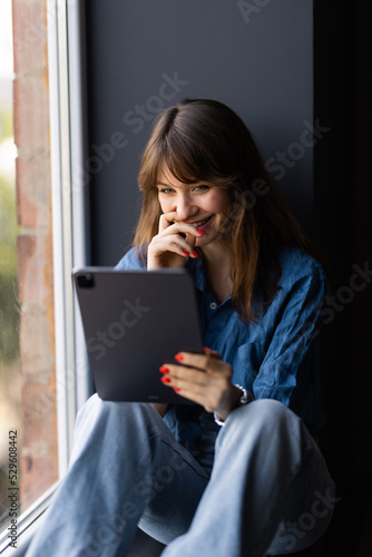 Portrait of positive woman with brown hair sitting on windowsill, smiling and holding tablet and white cup of tea in sunny morning.