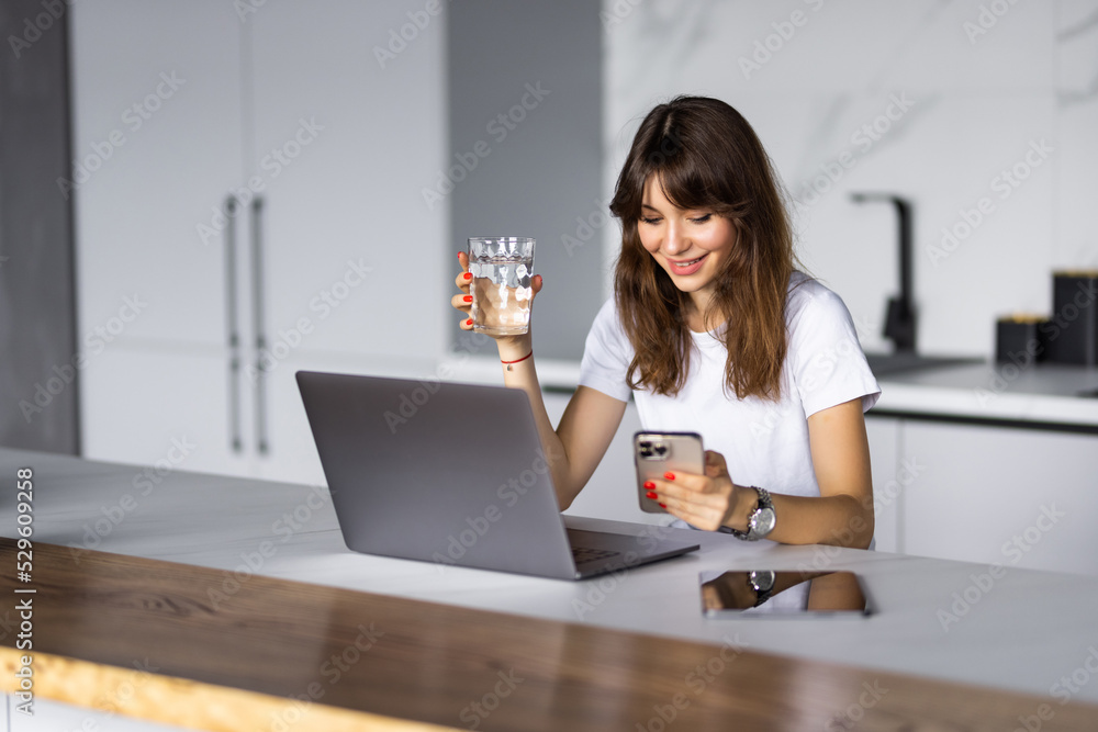 Smiling young woman using mobile phone while sitting on a kitchen with laptop Smiling young woman using mobile phone while sitting on a kitchen with laptop