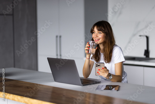 Smiling young woman using mobile phone while sitting on a kitchen with laptop