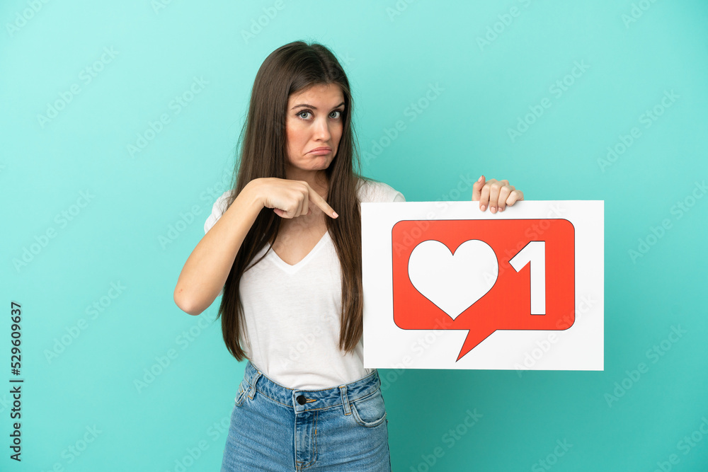 Young caucasian woman isolated on blue background holding a placard with Like icon and pointing it Young caucasian woman isolated on blue background holding a placard with Like icon and pointing it