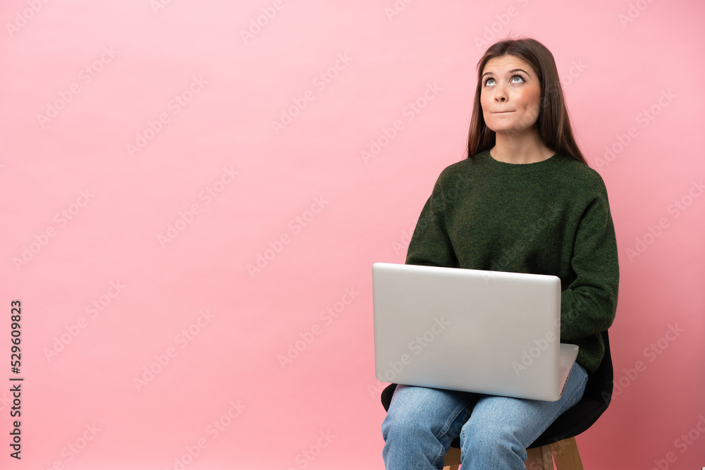 Naklejka premium Young caucasian woman sitting on a chair with her laptop isolated on pink background and looking up