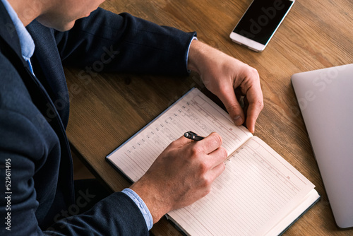 Young serious businessman writes something in notebook sitting at table with lying smartphone. Man wearing glasses fills notebook wit pen, organizing his affairs and life. Top view