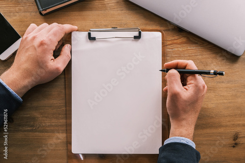 Business mockup, layout. Top view of clipboard with white sheet of paper vertically placed on wooden table. Male hands are holding pencil or ballpoint pen. Laptop, notepad and mobile phone lie nearby