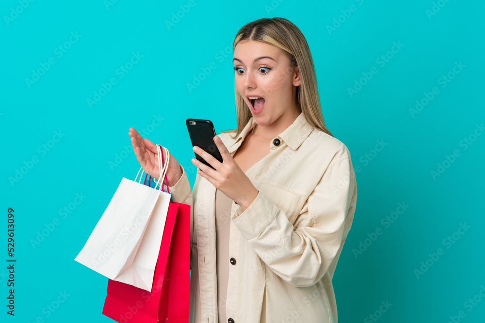 Young caucasian woman isolated on blue background holding shopping bags and writing a message with her cell phone to a friend Young caucasian woman isolated on blue background holding shopping bags and writing a message with her cell phone to a friend
