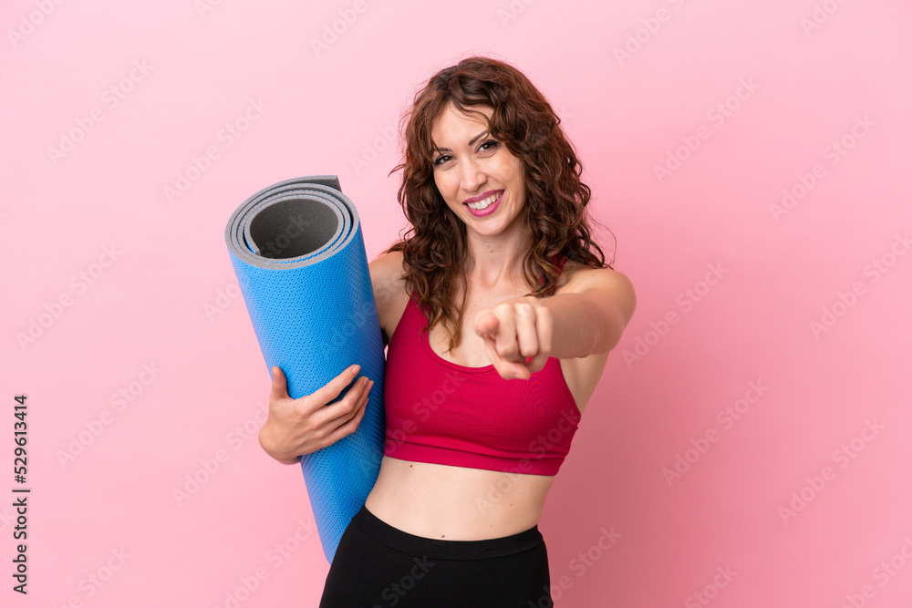 Young sport woman going to yoga classes while holding a mat isolated on pink background pointing front with happy expression Young sport woman going to yoga classes while holding a mat isolated on pink background pointing front with happy expression