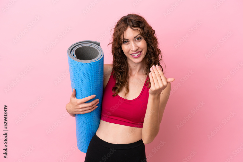 Young sport woman going to yoga classes while holding a mat isolated on pink background inviting to come with hand. Happy that you came Young sport woman going to yoga classes while holding a mat isolated on pink background inviting to come with hand. Happy that you came