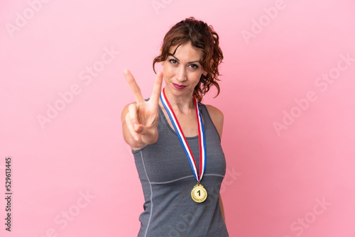 Young caucasian woman with medals isolated on pink background smiling and showing victory sign