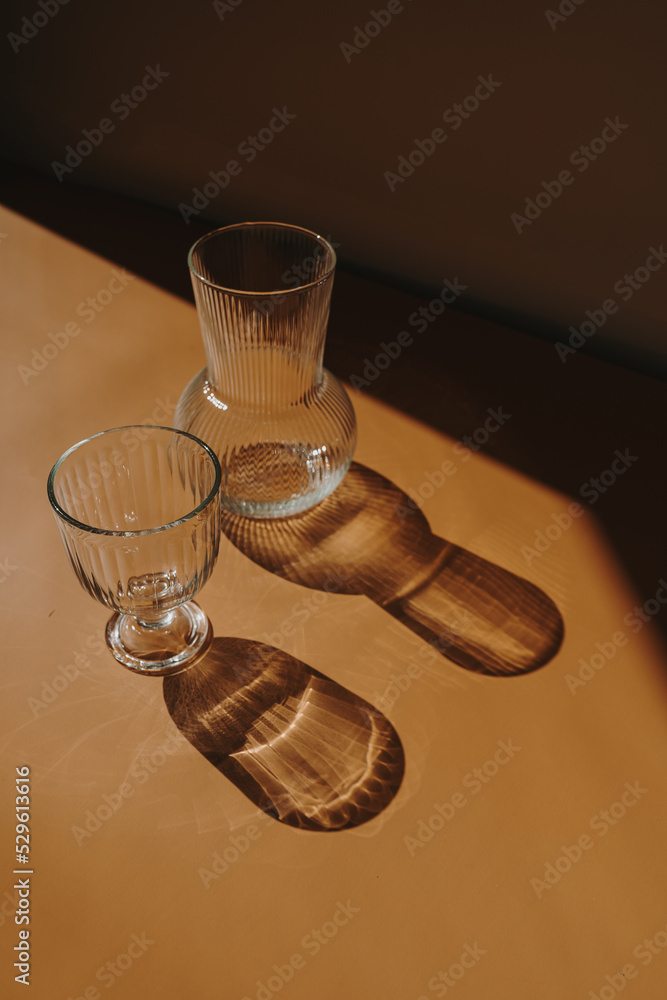 Glass and glass vase on orange background with sunlight shadows