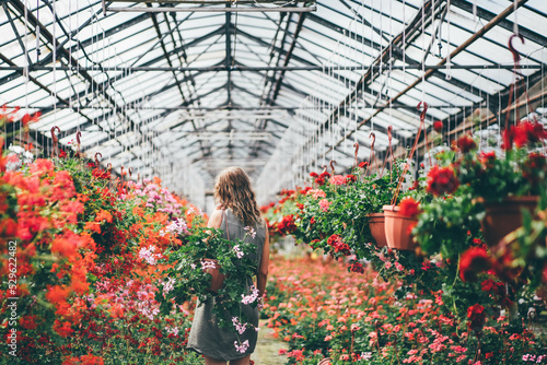 Happy young woman with fresh colorful flowers t walks along aromatic blooming plants in modern greenhouse on sunny day.
