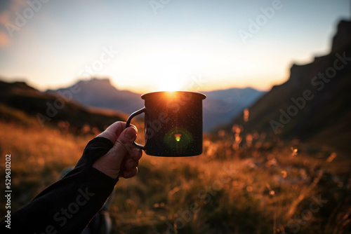 Closeup photo of cup with tea, coffee in traveler's hand over out of focus mountains view. Scenery in the Swiss mountains. Trekking concept