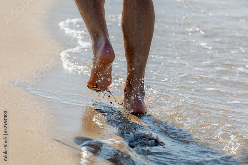 Keep walking concept, Selective focus of bare men's feet with approaching seawave on the shore, Low angle of of a man walking on the sand beach with footprints, Activities and recreation in summer.