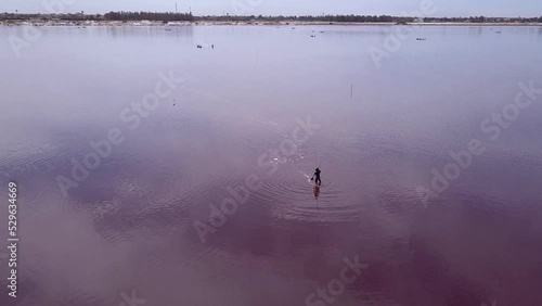 Aerial footage of man moving water collecting salt in middle of pink lake with small boat full of salt, clouds reflecting on the lake, Retba lake, Dakar, Senegal, going backward away from the man. photo