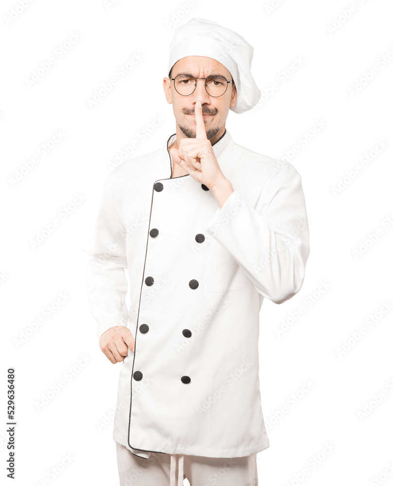 Serious young chef asking for silence gesturing with his finger Serious young chef asking for silence gesturing with his finger