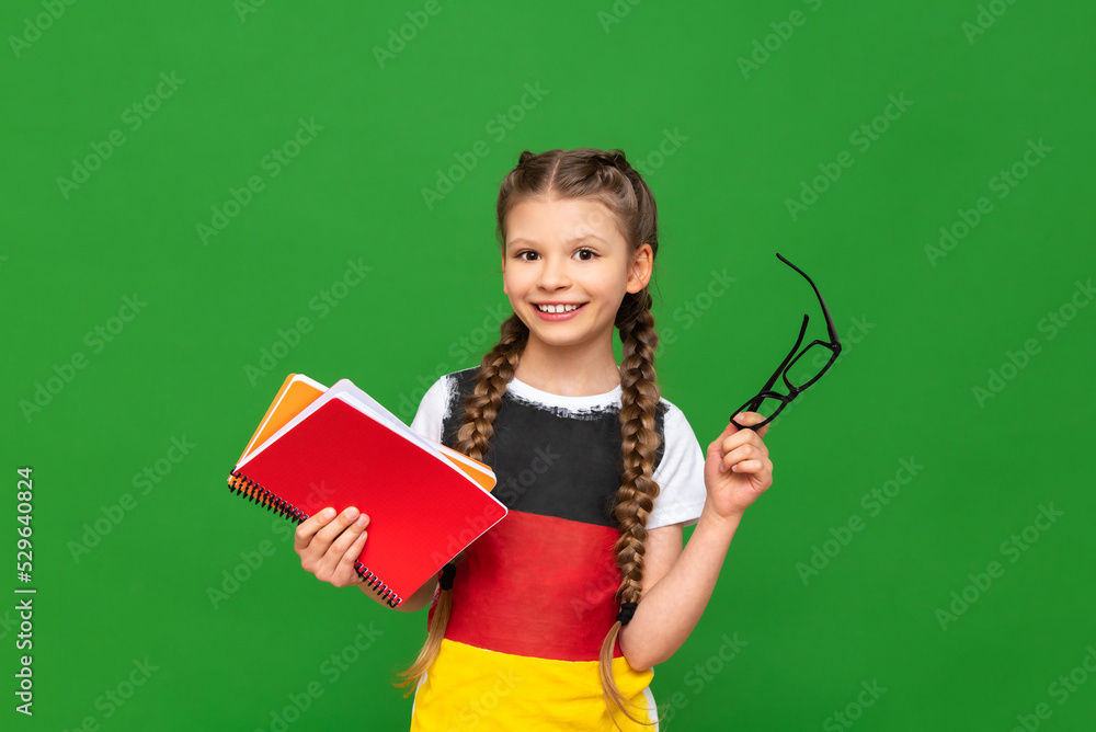 A child learns German on a green isolated background. A beautiful little girl with glasses with a German flag and notebooks on a green isolated background. A child learns German on a green isolated background. A beautiful little girl with glasses with a German flag and notebooks on a green isolated background.
