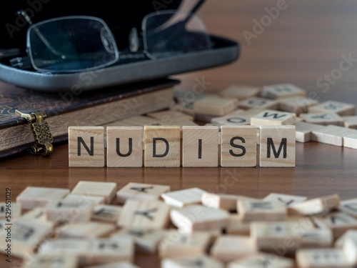 nudism word or concept represented by wooden letter tiles on a wooden table with glasses and a book