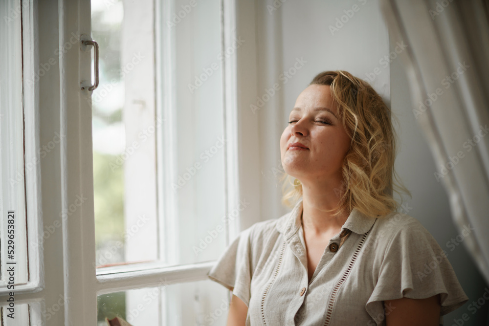 A middle-aged woman in a cream dress sits mysteriously and looks out the window on the windowsill. Green trees outside. A middle-aged woman in a cream dress sits mysteriously and looks out the window on the windowsill. Green trees outside.