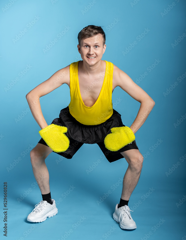 lean sportsman in a yellow t-shirt and black shorts on a blue background. funny jock put on yellow massage mittens and funny grimaces face lean sportsman in a yellow t-shirt and black shorts on a blue background. funny jock put on yellow massage mittens and funny grimaces face
