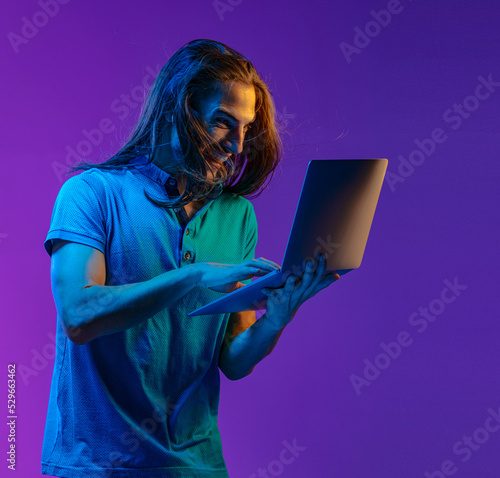 A nice emotional guy holds a laptop in his hands and looks at the screen with surprise. colored neon light
