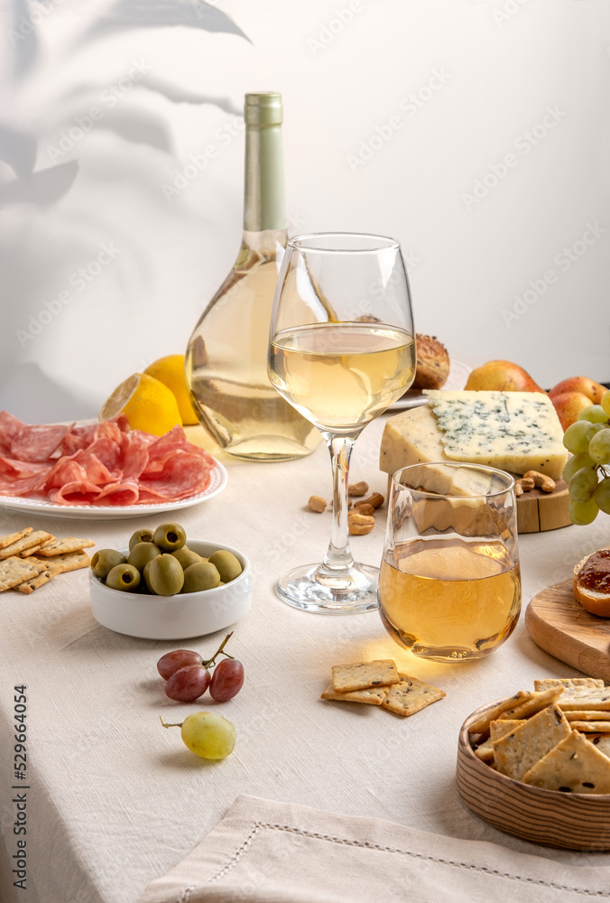 Glasses and bottle of white wine with grapes, cheese and salami on light table background. White wine with cheese and fruits. Wine bottle mockup with blank labels to place your design. Space for text