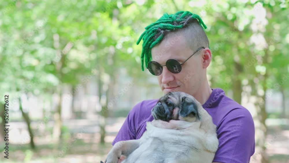 Hipster with green dreadlocks and black round glasses, holding and stroking a cute pug on a sunny summer day. A man walks his dog Hipster with green dreadlocks and black round glasses, holding and stroking a cute pug on a sunny summer day. A man walks his dog