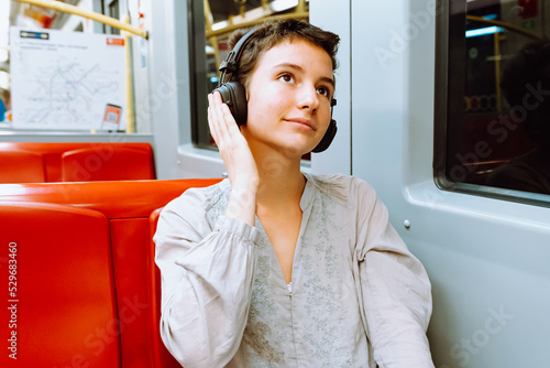 teenage girl with short haircut, smiling, listening to music in headphones while sitting in subway car or train