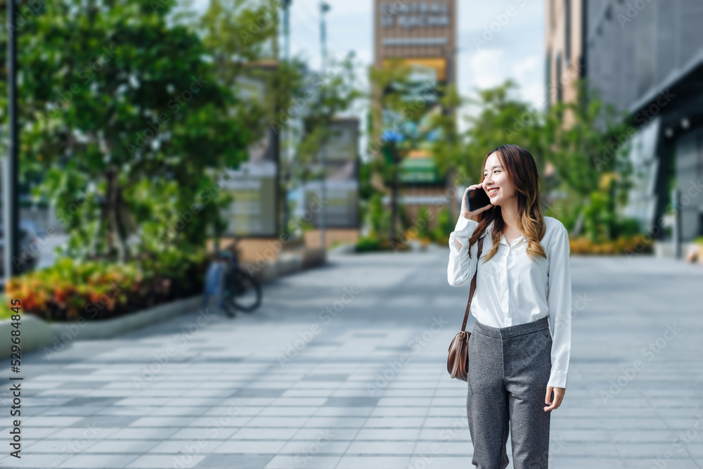 Fototapeta premium Smiling Asian curly business woman wearing trendy walks down the central city street and uses her phone. Pretty summer woman in white shirt walks down the street looking at her mobile phone