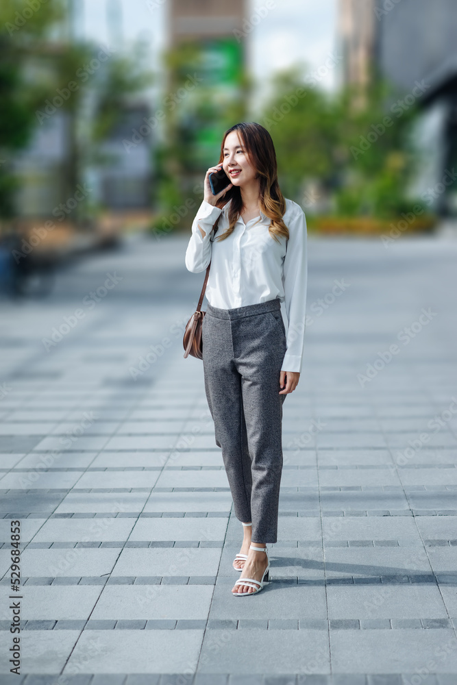 Smiling Asian curly business woman wearing trendy walks down the central city street and uses her phone. Pretty summer woman in white shirt walks down the street looking at her mobile phone Smiling Asian curly business woman wearing trendy walks down the central city street and uses her phone. Pretty summer woman in white shirt walks down the street looking at her mobile phone
