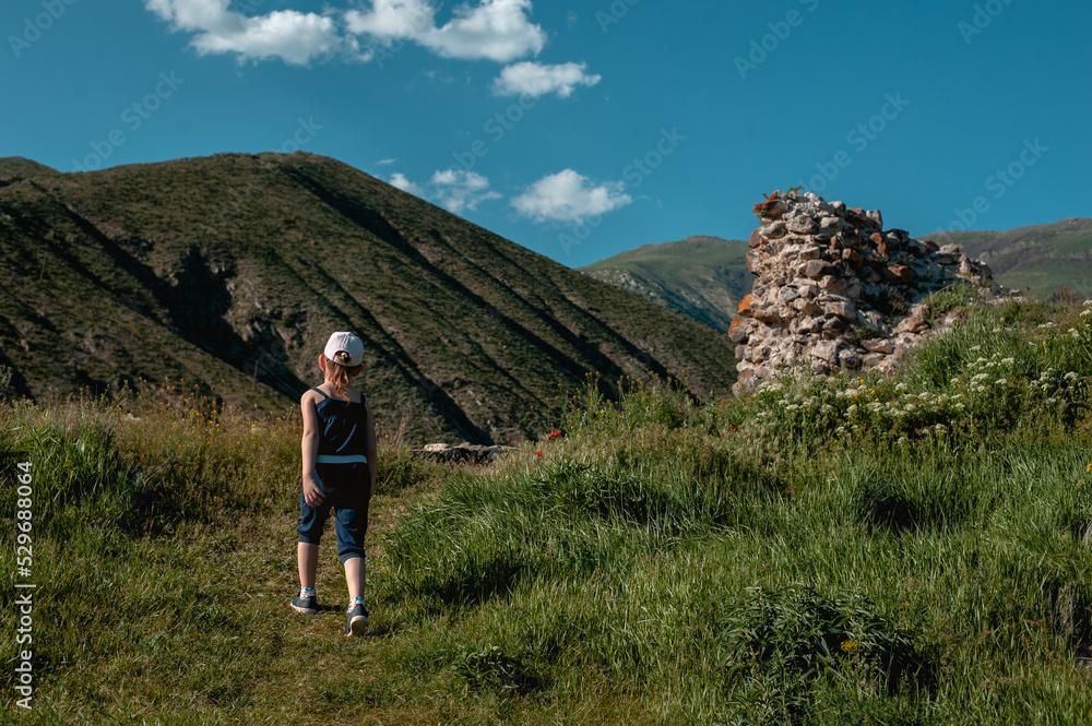 Fototapeta premium a preschooler girl in a cap walks alone in the mountains along a path, a view from the back. Little tourist on a hike in the mountains. Sky, mountains and a girl.