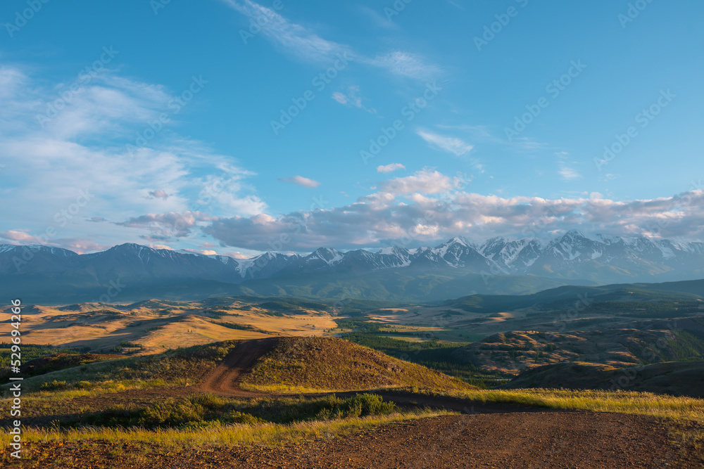 Scenic sunny view from sunlit grassy hill with dirt road to valley with hills and forest against high snowy mountain range in sunlight. Beautiful mountain landscape with snow mountains in sunshine.