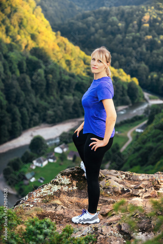 a blonde tourist woman stands on the very top of the mountain, a river flows below and small houses are visible