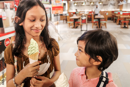 Southeast Asian children, boy and girl enjoy eating green tea and vanilla ice cream together at food court staring at each other looking happy