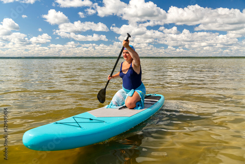 A Jewish woman in a kerchief kisuy rosh on her head and in a pareo on her knees on a SUP board with an oar swims in the lake on a sunny day. © finist_4