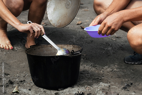 tourists, a man and a woman are pouring hot broth into plates, cooked outdoors on a fire in a large cast-iron pot