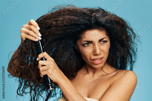 Print op canvas Angry upset curly Latin woman using a hair straightener, posing isolated on blue wall background, look aside