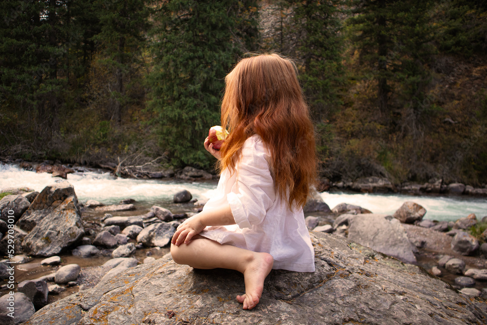 a little girl with long red hair in a white dress is sitting near the river and eating an apple a little girl with long red hair in a white dress is sitting near the river and eating an apple