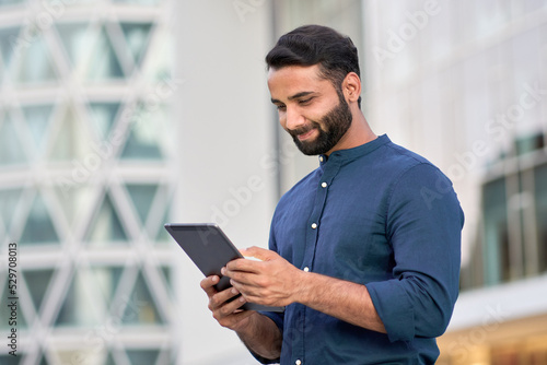 Murais de parede Smiling young adult indian business man professional, eastern businessman executive standing outdoors in urban city street checking data, holding using digital tablet online technology outside