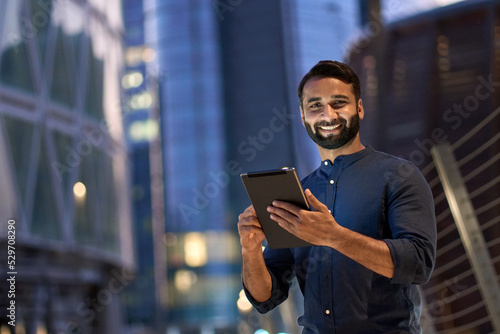 Smiling happy young eastern indian business man professional manager standing outdoor on street holding using digital tablet online fintech in night city with urban lights looking at camera, portrait. photo