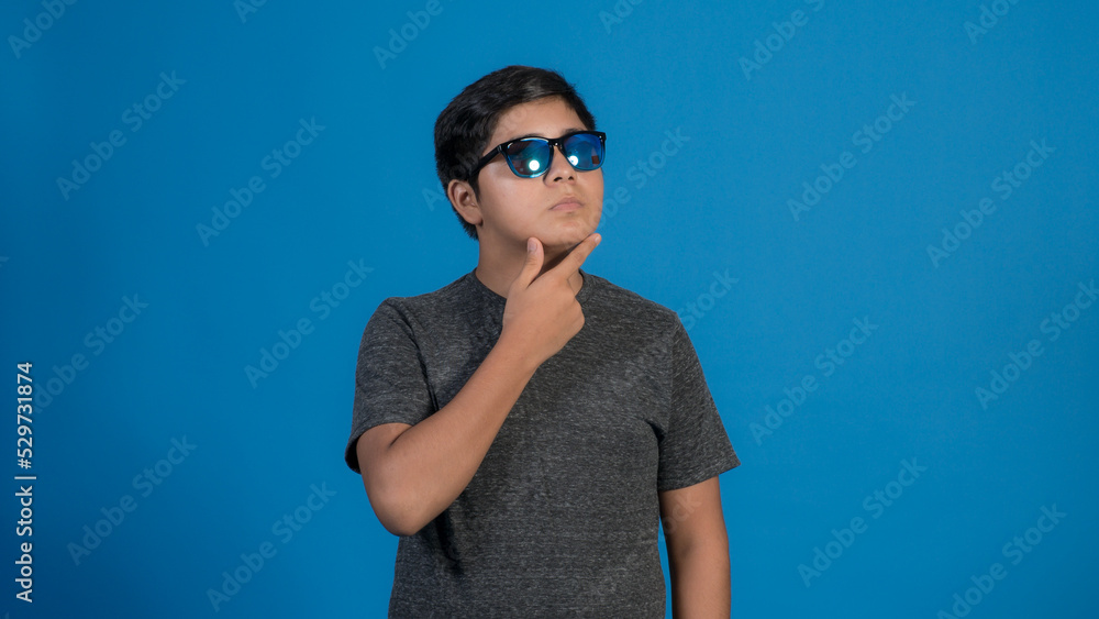 Foto de chico alegre llevar ropa de moda y lentes haciendo aislada en el fondo de color azul. Pensando tocando su cara con la mano.