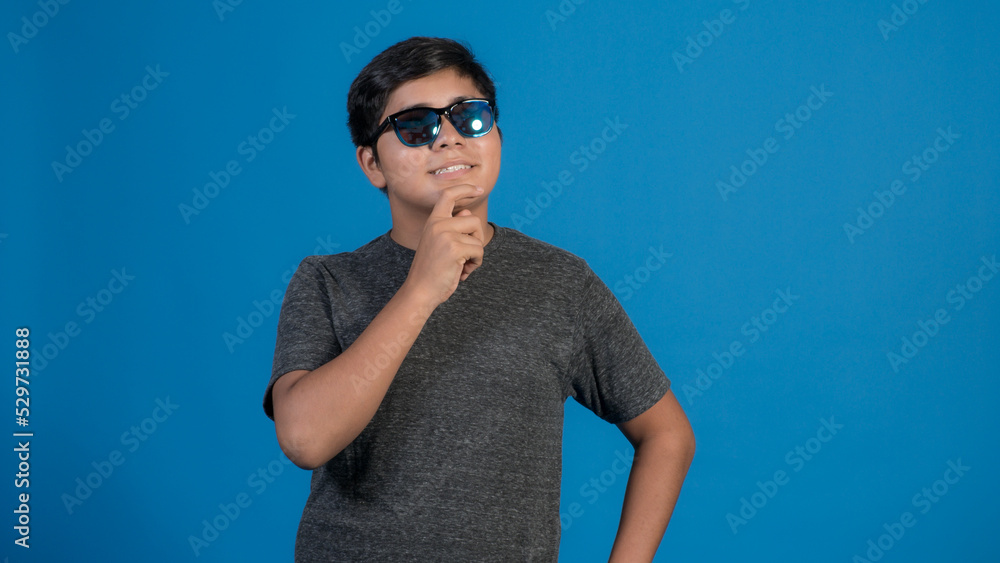 Foto de chico alegre llevar ropa de moda y lentes haciendo aislada en el fondo de color azul. Pensando tocando su cara con la mano. Foto de chico alegre llevar ropa de moda y lentes haciendo aislada en el fondo de color azul. Pensando tocando su cara con la mano.
