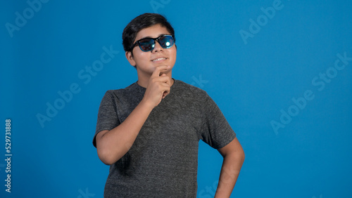 Foto de chico alegre llevar ropa de moda y lentes haciendo aislada en el fondo de color azul. Pensando tocando su cara con la mano.