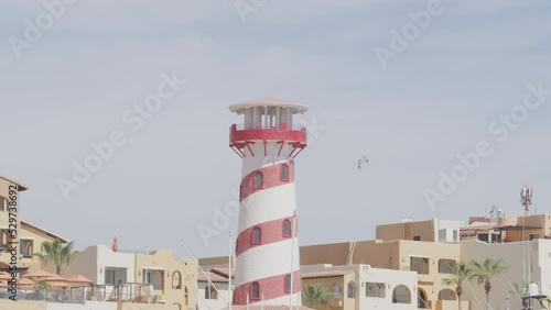 Seagull flying past red and white striped lighthouse in Cabo San Lucas, Mexico with resorts and apartments in background photo