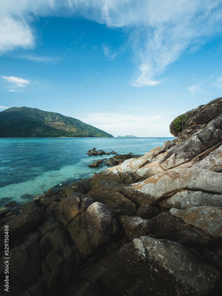 Scenic view of Koh Kra, peaceful small island rocky coast and clear turquoise sea with coral reef transparent against summer blue sky and Koh Adang Island background. Near Koh Lipe, Satun, Thailand.