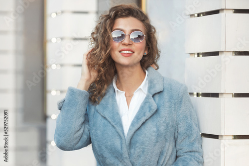 Portrait of happy millennial woman 30-35 years old with curly hair in blue trendy sunglasses and faux fur coat and white shirt photo