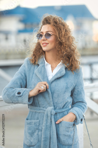 Lifestyle curly beautiful woman in blue autumn fur coat and colored sunglasses walking in the city