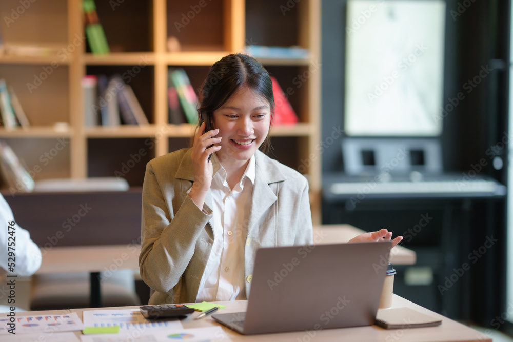 Young Asian businesswoman sit with her laptop computer happily talking on the phone with her customer explaining the detail in the office. Young Asian businesswoman sit with her laptop computer happily talking on the phone with her customer explaining the detail in the office.