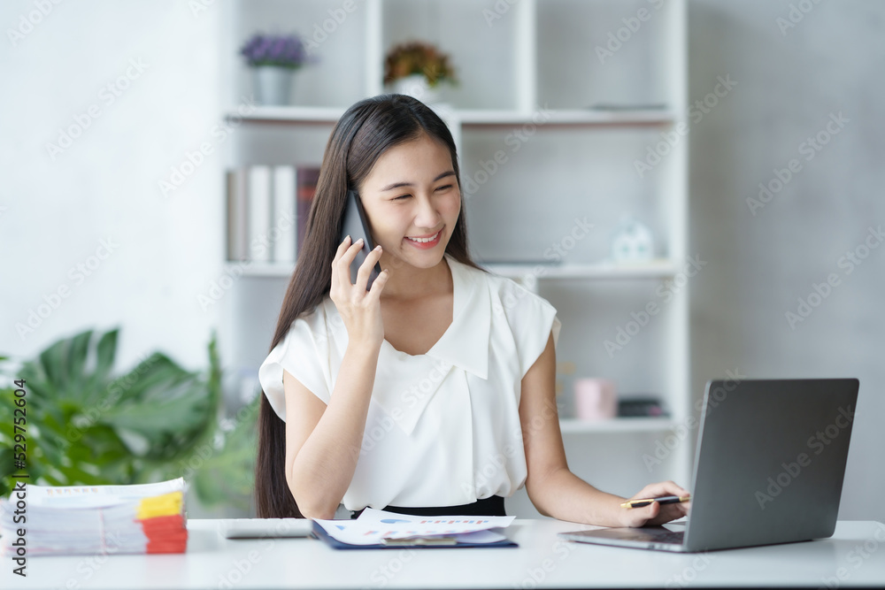 Young Asian businesswoman sit with her laptop computer happily talking on the phone with her customer explaining the detail in the office. Young Asian businesswoman sit with her laptop computer happily talking on the phone with her customer explaining the detail in the office.