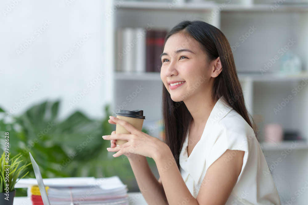 At the office, a young Asian businesswoman sit and relaxe with a cup of coffee and a bright smile on her laptop. At the office, a young Asian businesswoman sit and relaxe with a cup of coffee and a bright smile on her laptop.